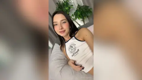 Smiling young woman in a white graphic tank top relaxing on a bed in a bright, cozy room.