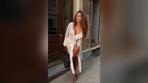 InesMoore poses outdoors in a long white ruffled dress with a high slit, showing her long legs and wearing strappy heels.