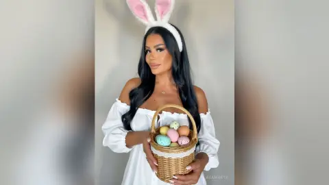 Smiling woman in white off-the-shoulder dress holding a basket of colorful Easter eggs with bunny ears.