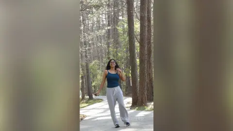 Smiling woman in a blue tank top and grey sweatpants walking through a sunny, lush pine forest.