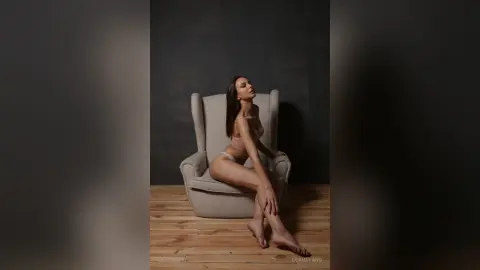 LeahEvans sits on a grey wingback chair wearing light pink lingerie against a dark background.