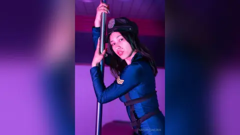 NashiroSakurai poses while holding a silver pole, wearing a blue police-style uniform and cap.