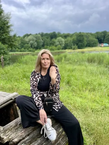 NikaZen sits on a wooden log in a grassy field wearing a leopard print shirt and black pants.