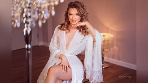 Beautiful woman in a white lace robe sitting in a warm, elegant room with soft lighting.
