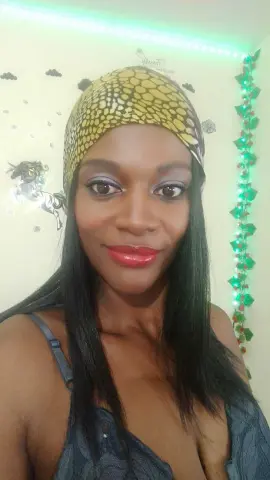 Smiling Black woman in a yellow patterned headwrap and grey tank top posing indoors with festive lights.