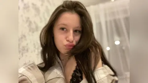 Young woman with long brown hair wearing a cozy cream sweater in a bright, airy room.