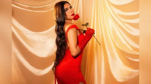 Elegant woman in a red cocktail dress holding a single rose against a luxurious gold draped backdrop.