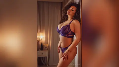 Curvy woman in purple lace lingerie posing confidently in a warm, dimly lit bedroom setting.