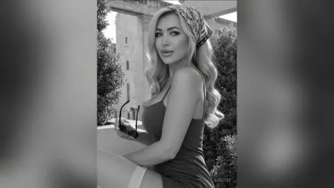 Anelise poses sideways in a dark tank top and headscarf, showing her blonde wavy hair and curves against a stone building and greenery.
