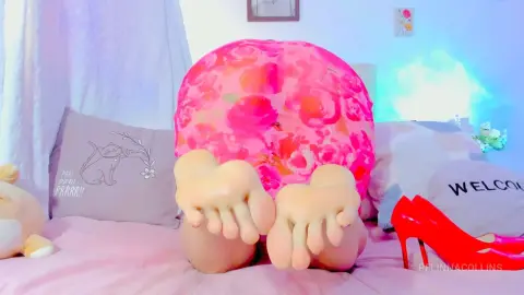PelinnaCollins lying on a pink bed wearing a pink floral top, showing her bare feet close up to the camera.