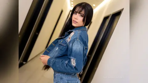 Young woman in a distressed denim jacket posing in a modern minimalist hallway with soft lighting.