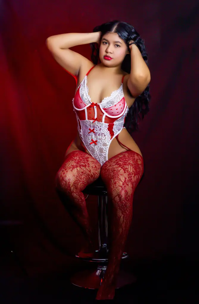 NataliEvansx — NataliEvansx poses in a red and white lace bodysuit and red lace stockings against a dark red background.