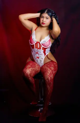 NataliEvansx poses in a red and white lace bodysuit and red lace stockings against a dark red background.
