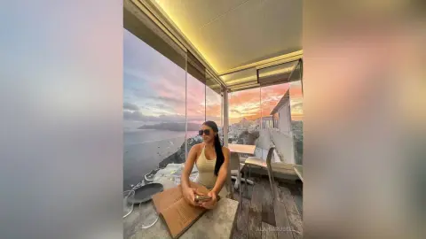 Stylish woman in a beige tank top reading a book on a luxury balcony overlooking a scenic ocean sunset.