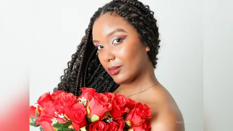 Beautiful Black woman with long braids holding a red rose bouquet in a bright studio setting.