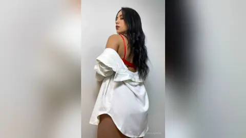Young woman in a white oversized shirt and red bra posing against a plain wall for a stylish portrait.