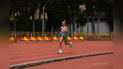 Athletic woman in green sports bra and shorts running on a red outdoor track under bright daylight.