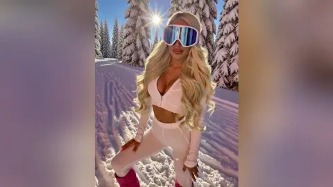 Blonde woman in white ski outfit and goggles posing in a sunny snowy mountain landscape.