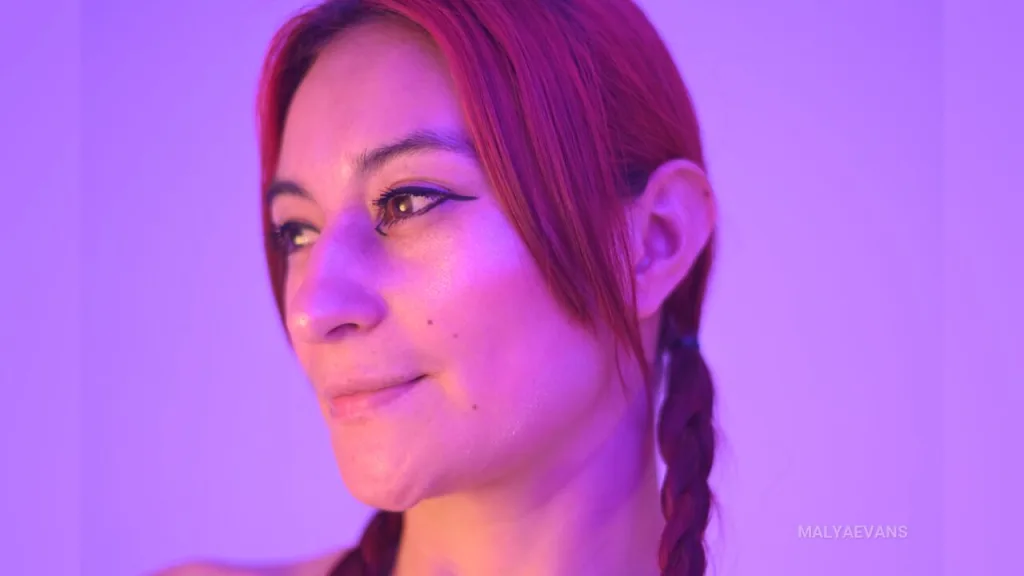 MalyaEvans poses with her hands pulling down a red lace bra to expose her breasts against a purple background.