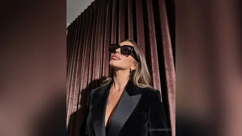 Stylish woman in a black blazer and sunglasses posing confidently against elegant velvet curtains.