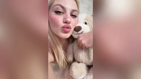 Blonde woman in a white top blowing a kiss while hugging a large beige teddy bear in a cozy indoor setting.