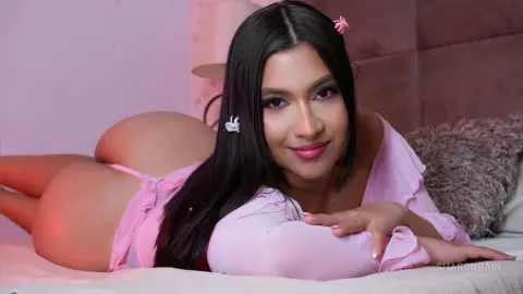 SharonRain lies on a bed in a pink long-sleeve top, showing her bare buttocks and curves while looking at the camera with long dark hair.