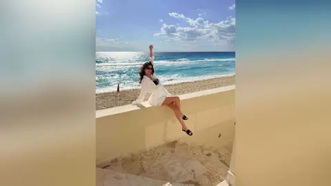 Cheerful woman in a white breezy outfit sitting on a beach wall under a sunny blue sky.