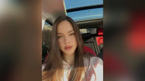 Young woman with long brown hair wearing a beige top sitting in a car under bright sunlight.