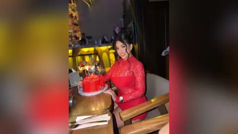 Smiling woman in a red lace dress celebrating a birthday with a lit cake in a dimly lit restaurant.