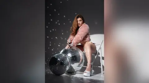 Stylish woman in a pink faux fur coat sitting by disco balls in a glamorous studio setting.