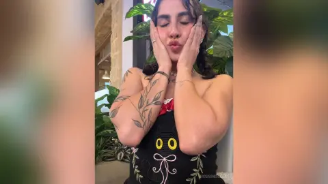 Young woman in a black graphic tank top posing playfully with hands on cheeks in a lush indoor garden setting.
