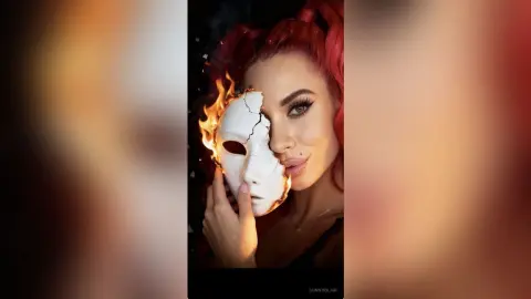 Red-haired woman holding a white cracked mask with flames in a dramatic, mystical setting.