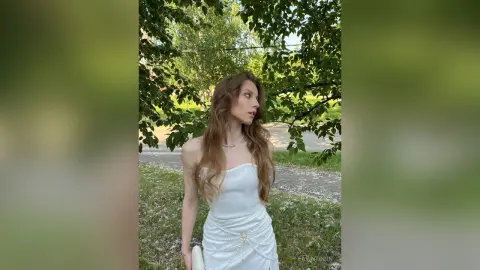 Elegant woman in a white satin slip dress posing in a lush green garden setting with a serene mood.