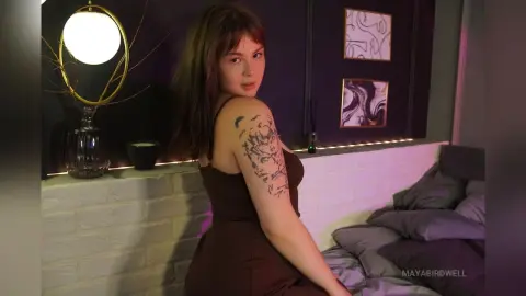 Young woman in a brown sleeveless dress with arm tattoos sitting in a moody, dimly lit modern bedroom.