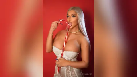 Blonde woman in white lace lingerie licking a candy cane against a vibrant red background.