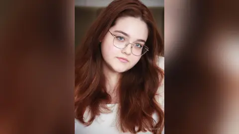 Portrait of a young woman with long red hair and gold glasses wearing a white top in a soft, natural light setting.