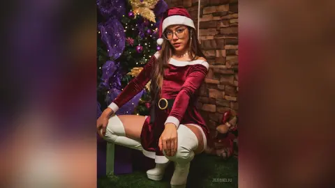 JessieLiu wears a red velvet off-the-shoulder Christmas dress and white thigh-high boots while posing next to a decorated tree.
