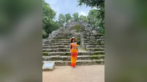 NohomiCambel stands in front of a stone pyramid wearing an orange bikini top and a long orange skirt with a wide-brimmed straw hat.