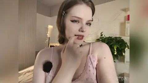 Young woman with winged eyeliner in a pink lace camisole posing in a bright, modern bedroom.