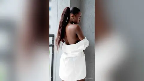 Elegant woman in a white off-the-shoulder dress posing gracefully against a minimalist wall background.