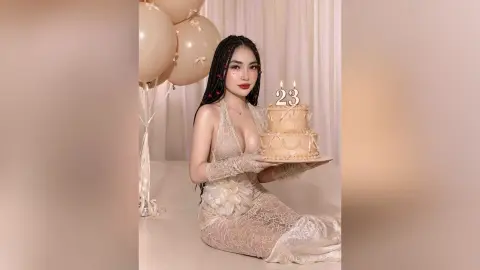 Young woman in a lace cream dress holding a 23rd birthday cake with balloons in a soft aesthetic setting.