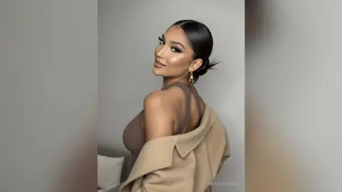 Glamorous woman in a brown ribbed top and beige coat with sleek hair and glowing makeup in a neutral studio.
