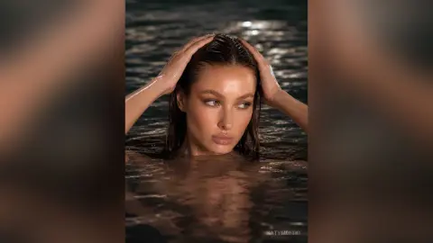 KatyMonro poses in dark water with her wet hair pulled back, looking directly at the camera.