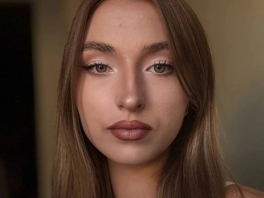 Close-up portrait of a young woman with natural makeup and long brown hair in soft indoor lighting.