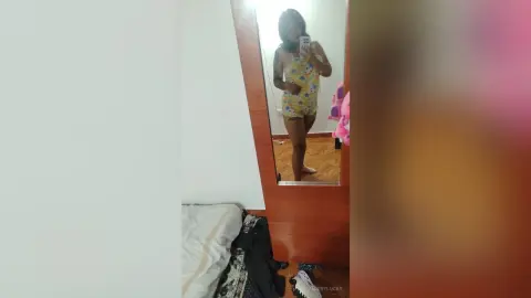 Young woman in a yellow floral romper taking a mirror selfie in a bright bedroom.