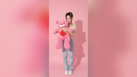 Young woman in a white crop top and light blue jeans hugging a large pink teddy bear against a pink background.