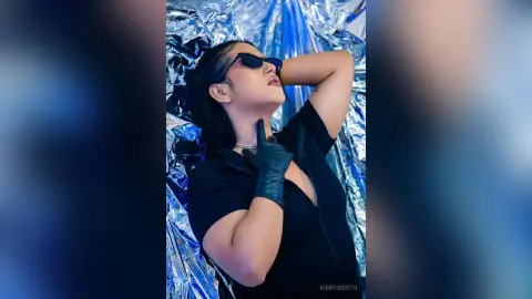 KimRoberth poses against a shiny silver foil background wearing a black zip-up top, black gloves, and sunglasses.