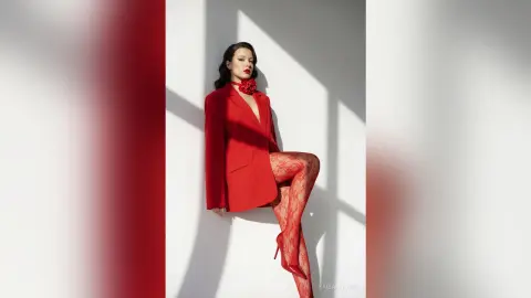 Elegant woman in a red blazer and patterned tights posing in bright sunlight against a white wall.