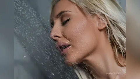 Anita has wet blonde hair and glistening skin while standing under a spray of water droplets.