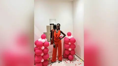 Stylish woman in a vibrant red jumpsuit posing next to pink balloon decor in a modern room.